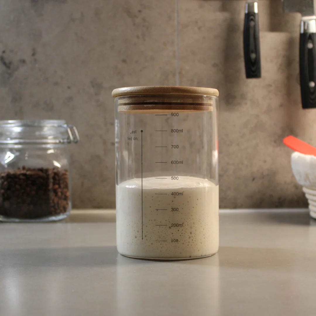 Surdeg i glasburk på en köksbänk En glasburk med surdeg uppmätt i milliliter med trälock. I bakgrunden syns en glasburk med kaffebönor, två svarta knivhandtag samt en jäskorg med en röd slev på en modern köksbänk.