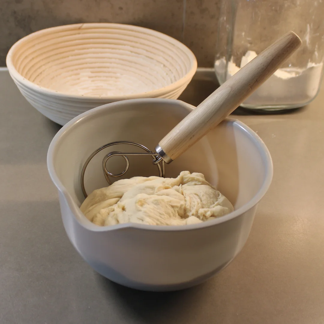 dansk_degvisp_25cm_3 En grå skål med deg och en dansk degvisp med trähandtag. I bakgrunden finns en jäskorg av rotting och en glasburk med mjöl på en grå köksbänk