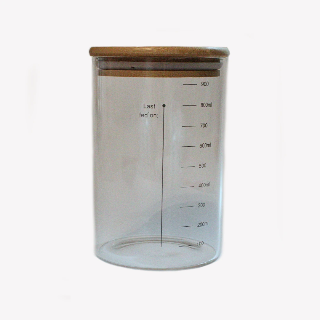 glasburk_1000ml_bred En bred glasburk med trälock för surdegstarter. Glasburken har tydliga mått linjer. Glasburken står på en vit bakgrund.