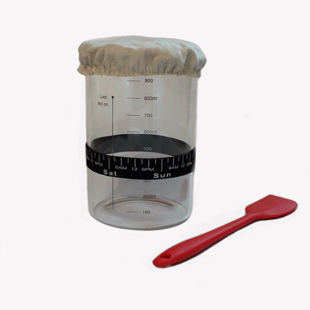 glasburk_1000ml_bred_2_new Startkit för surdeg. Kitet innehåller en bred glasburk med trälock för surdegstarter. Glasburken har tydliga måttangivelser, ett elastiskt tygöverdrag på toppen och en svart rund remsa runt burken markerad med tider och dagar för att hålla koll på surdegens matningar. En röd slickepott i silikon ligger bredvid burken. Burken står på en ljusgrå bakgrund.