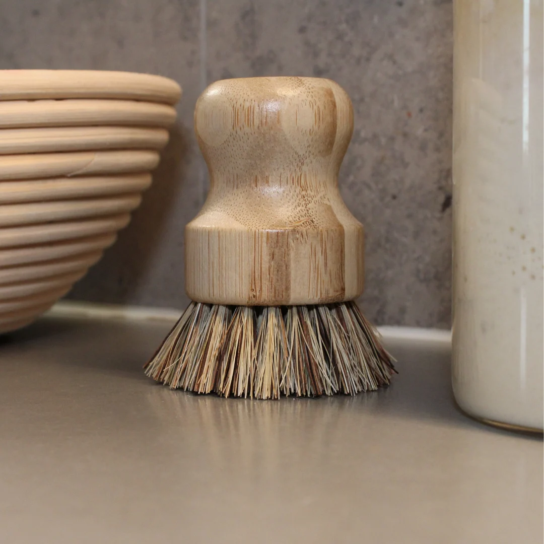 jaskorgsborste_2 En jäskorgsborste med trähandtag och naturborst står på en köksbänk. I bakgrunden syns en jäskorg, en glasburk och en grå marmorvägg.