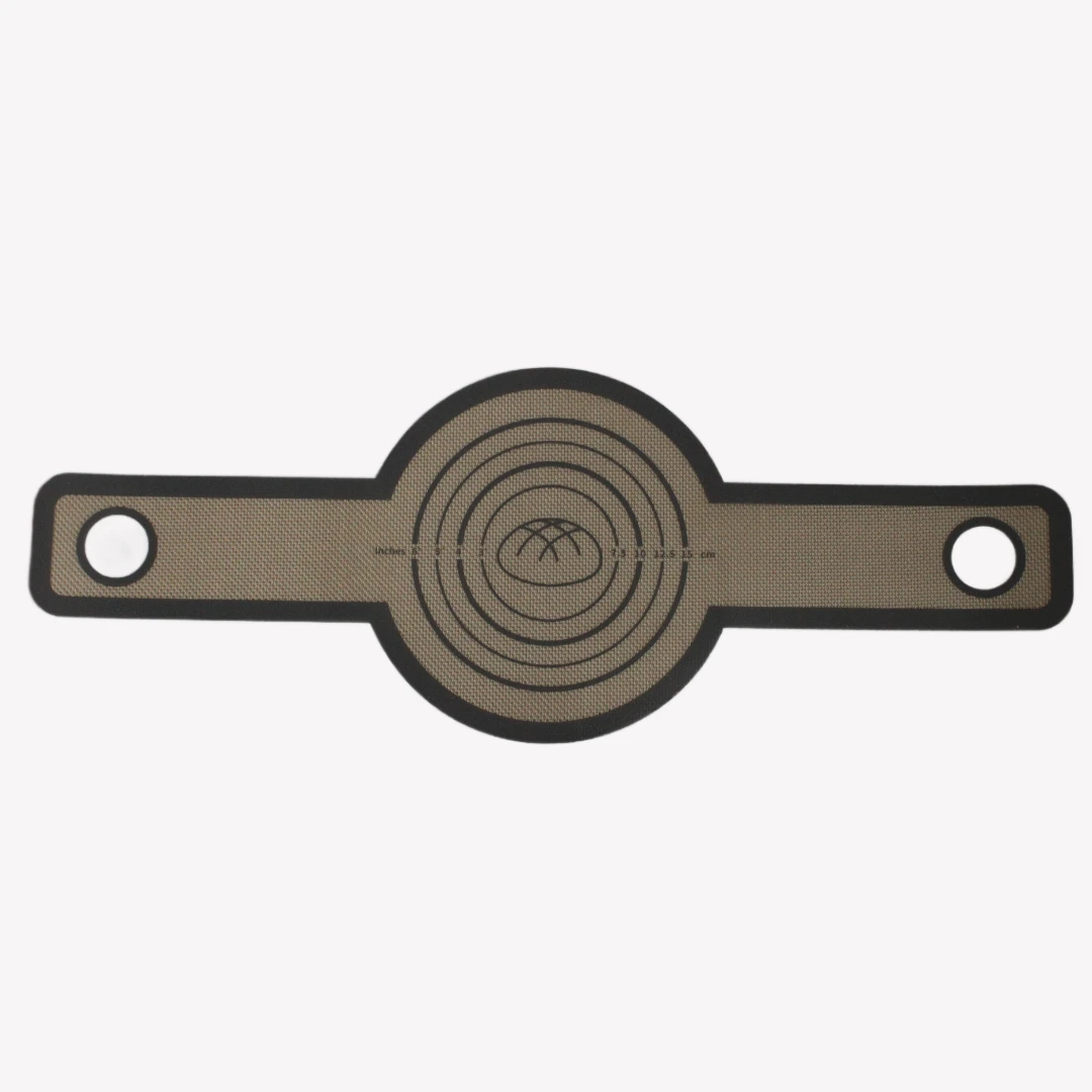 En beige silikonbakmatta för gjutjärnsgryta med svarttryckta cirklar och handtag med hål. I mitten av bakmattan finns en illustration av en brödlimpa. Bakmattan är utrullad på en vit bakgrund.