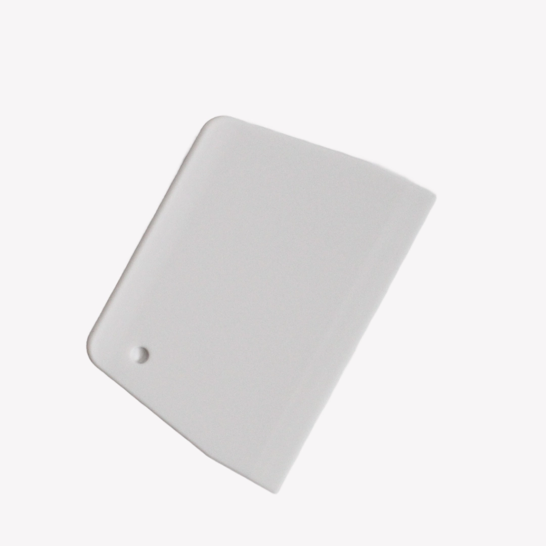 En vit plast degskrapa med rundade hörn ligger på en vit bakgrund. I det nedre vänstra hörnet av degskrapan finns ett runt hål.