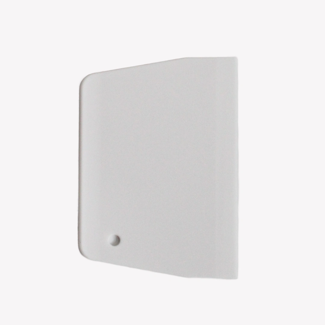 En vit plast degskrapa med rundade hörn ligger på en vit bakgrund. I det nedre vänstra hörnet av degskrapan finns ett runt hål.