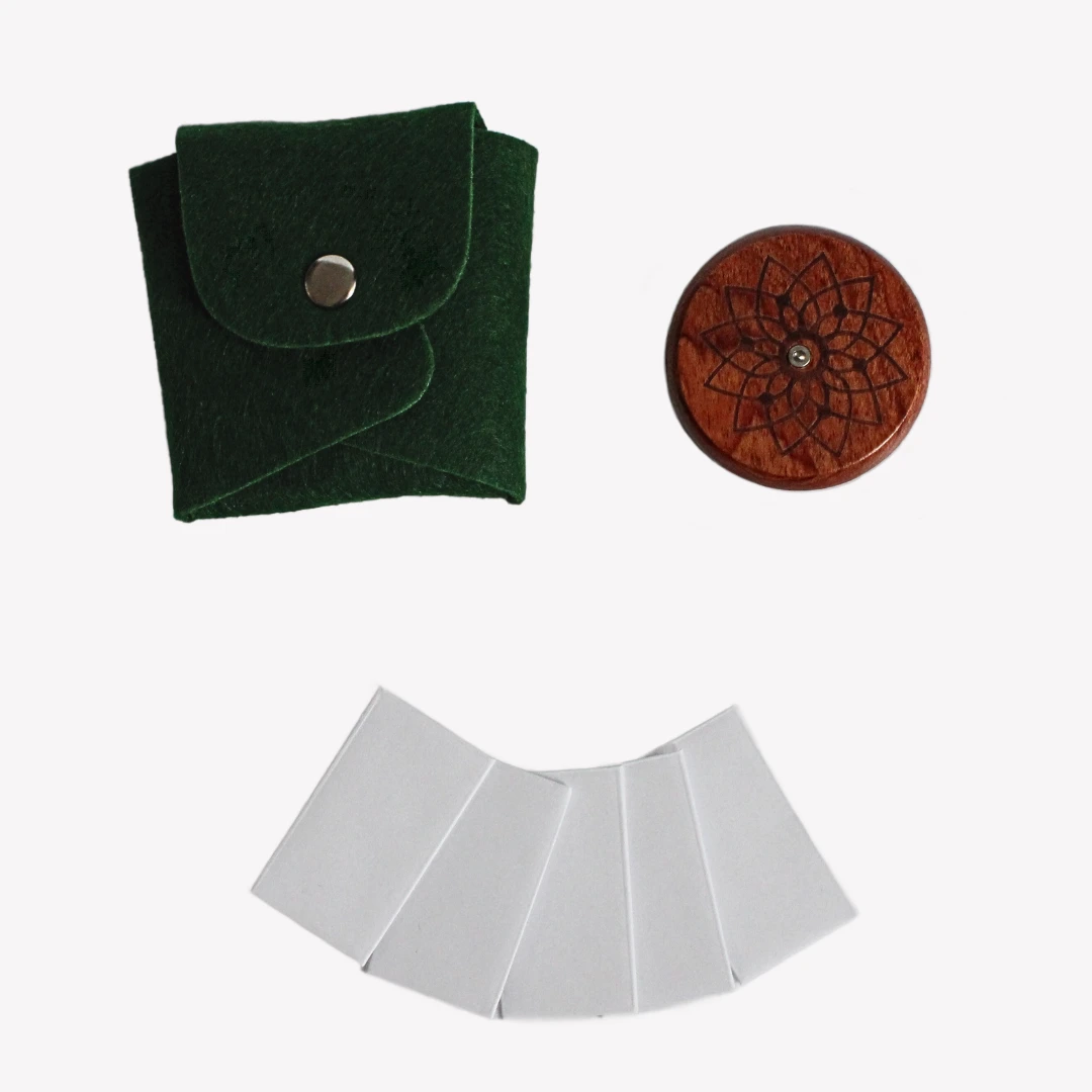 En rund snittkniv i trä med ett ingraverat mönster, liggande bredvid ett mörkgrönt fodral i läderimitation och fem metallblad inslagna i papper. Allt är placerat på en vit bakgrund.