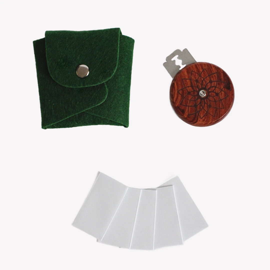 En rund snittkniv i trä med ett ingraverat mönster, liggande bredvid ett mörkgrönt fodral i läderimitation och fem metallblad inslagna i papper. Allt är placerat på en vit bakgrund.