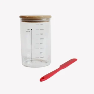 En bred glasburk med trälock för surdegstarter med tydliga måttlinjer. Bredvid glasburken ligger en röd slickepott i silikon. Bakgrunden är vit.