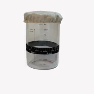 Startkit för surdeg. Kitet innehåller en bred glasburk med trälock för surdegstarter. Glasburken har tydliga måttangivelser, ett elastiskt tygöverdrag på toppen och en svart rund remsa runt burken markerad med tider och dagar för att hålla koll på surdegens matningar. Burken står på en ljusgrå bakgrund.